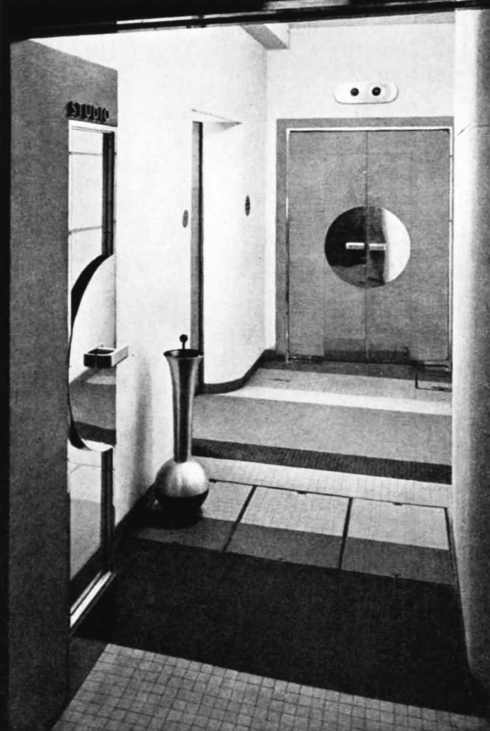 A corridor with a fancy free-standing ashtray, leading to wooden doors with a shiny metal circular push-plate