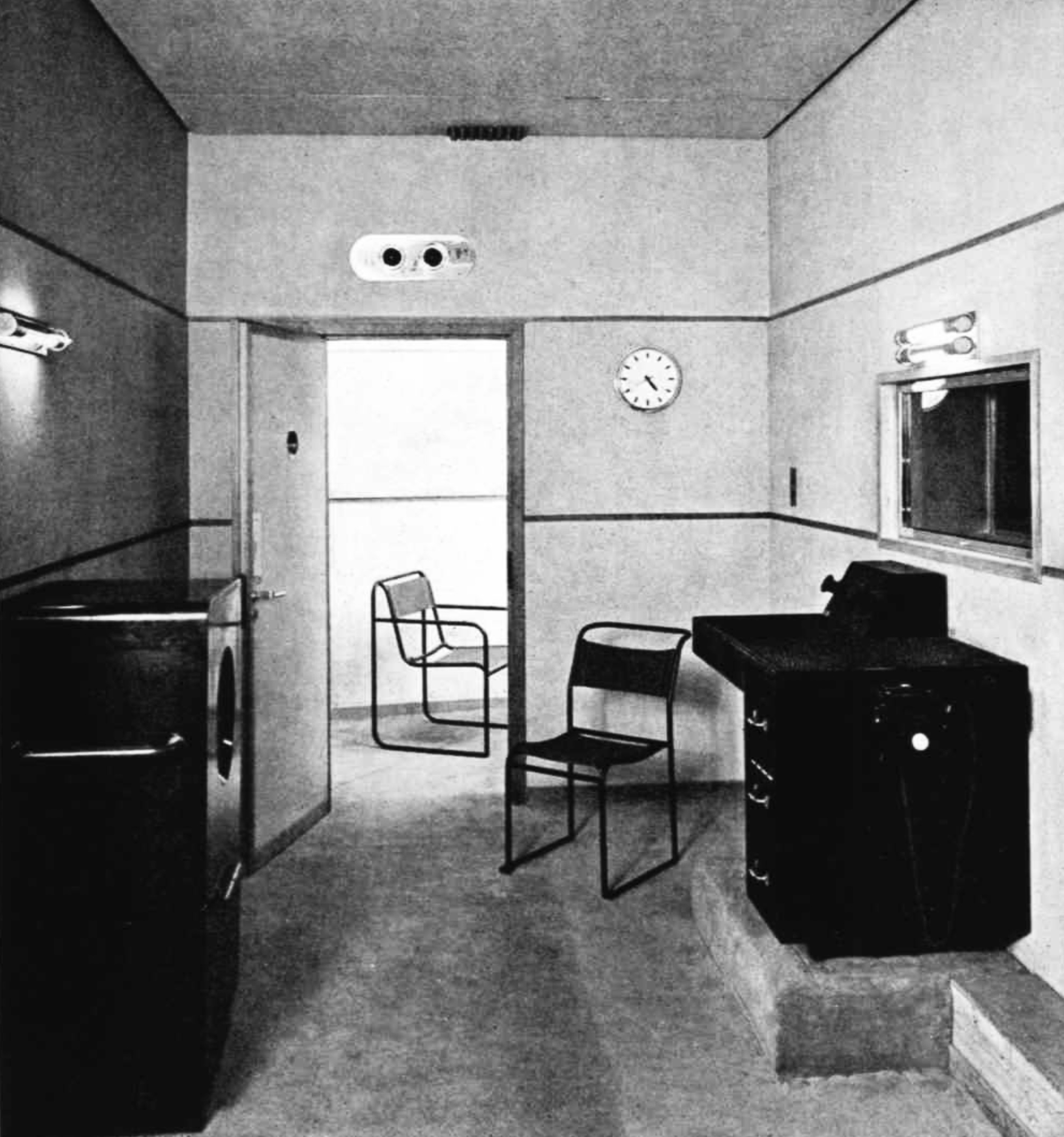 A control desk and a large speaker with an internal window to a studio; a door stands open with a single chair in the room beyond