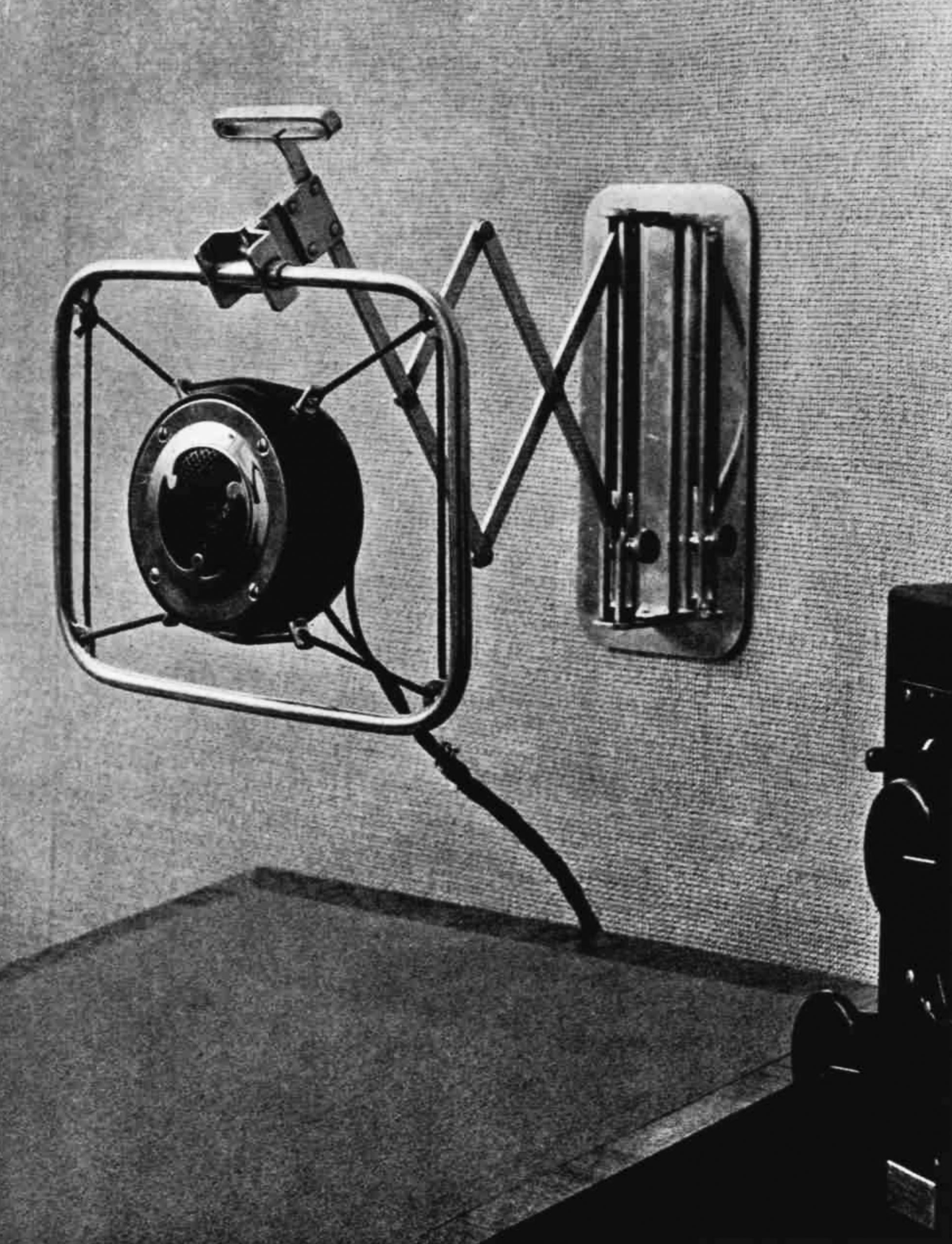A round microphone in a square frame, bouncing from the wall on a scissor-like projection