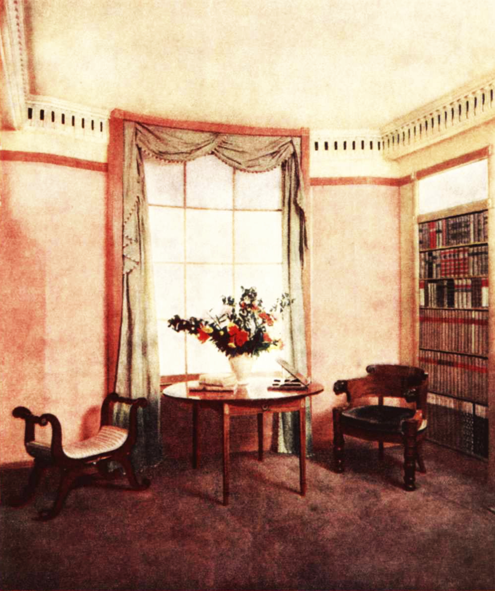 A colour picture of a cozy carpeted room with a bookcase, a curtained window, a round table and two easy chairs