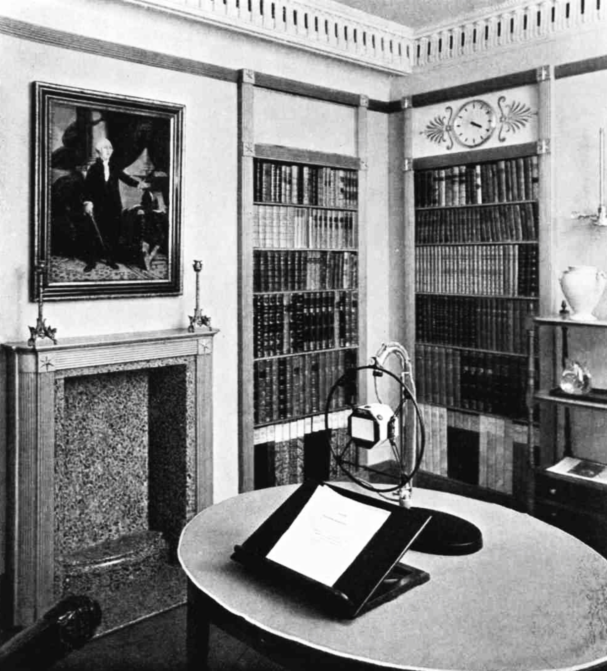 A room with a dummy fireplace, bookcases, ornaments and a round table with a microphone and a book stand