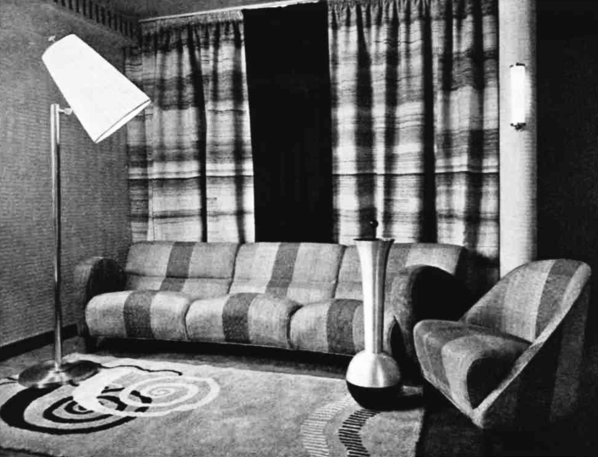 A curtained room with a rug, a sofa, a chair, a standard lamp and a free-standing ashtray